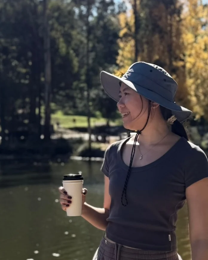 Ran Fang by a lake on a sunny autumn day, smiling and holding a coffee